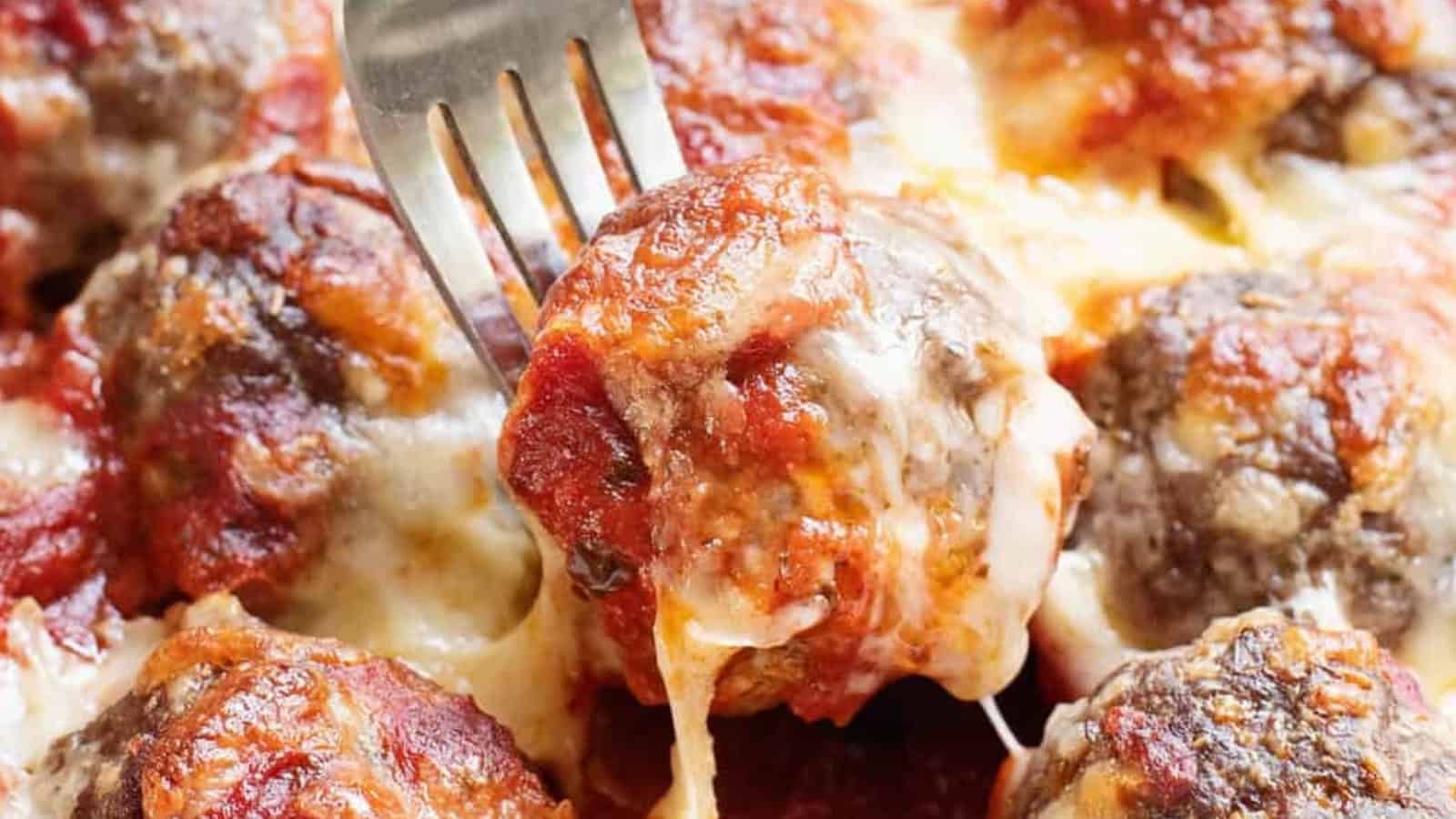 A fork lifting a cheesy, saucy meatball from a tray of baked meatballs topped with melted cheese.