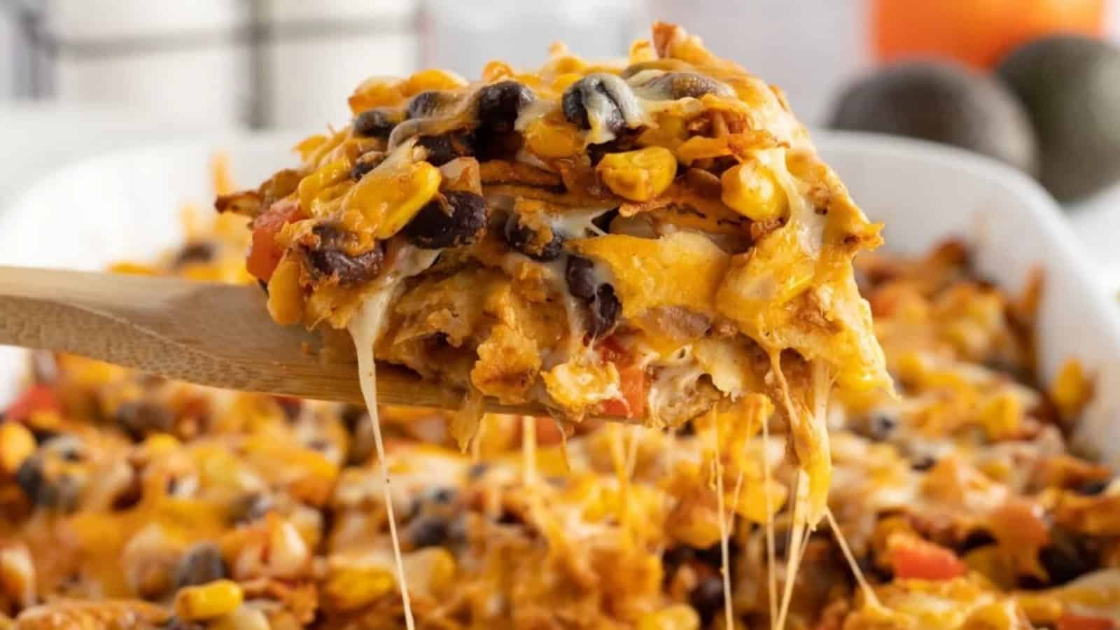 A wooden spoon lifts a cheesy, baked Mexican casserole with beans, corn, and melted cheese.