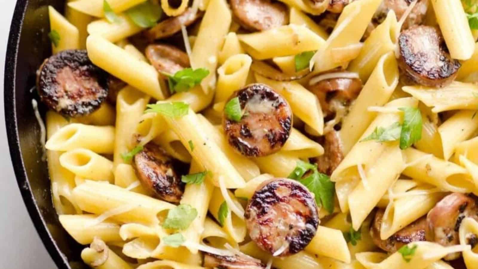 Penne pasta with browned sausage slices, mushrooms, and fresh parsley in a skillet.