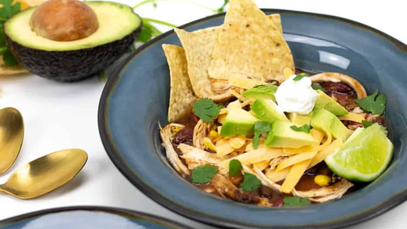 A bowl of tortilla soup with avocado, lime, chips, sour cream, and cilantro, next to a halved avocado.