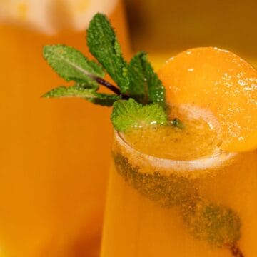A peach-colored drink garnished with mint leaves and a peach slice in a glass on a yellow surface.