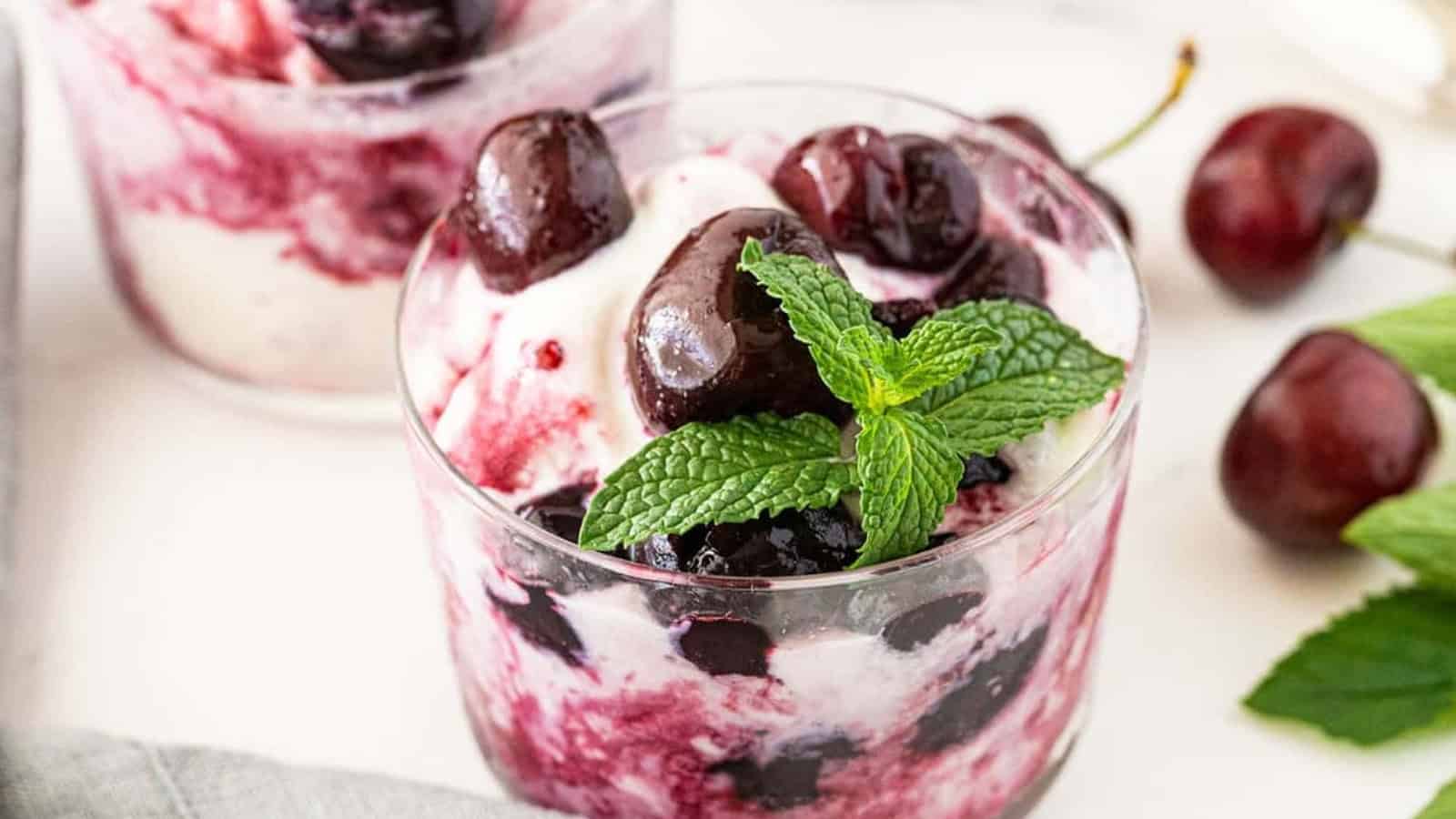 A glass of cherry parfait with swirls of cream, fresh cherries, and a sprig of mint on top.