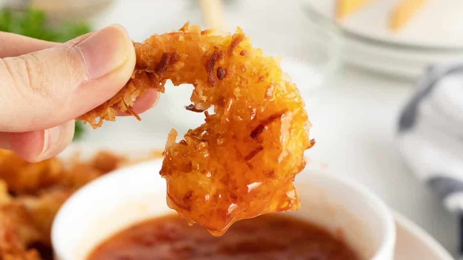 A hand holds a crispy coconut shrimp dipped in a red sauce over a white bowl.