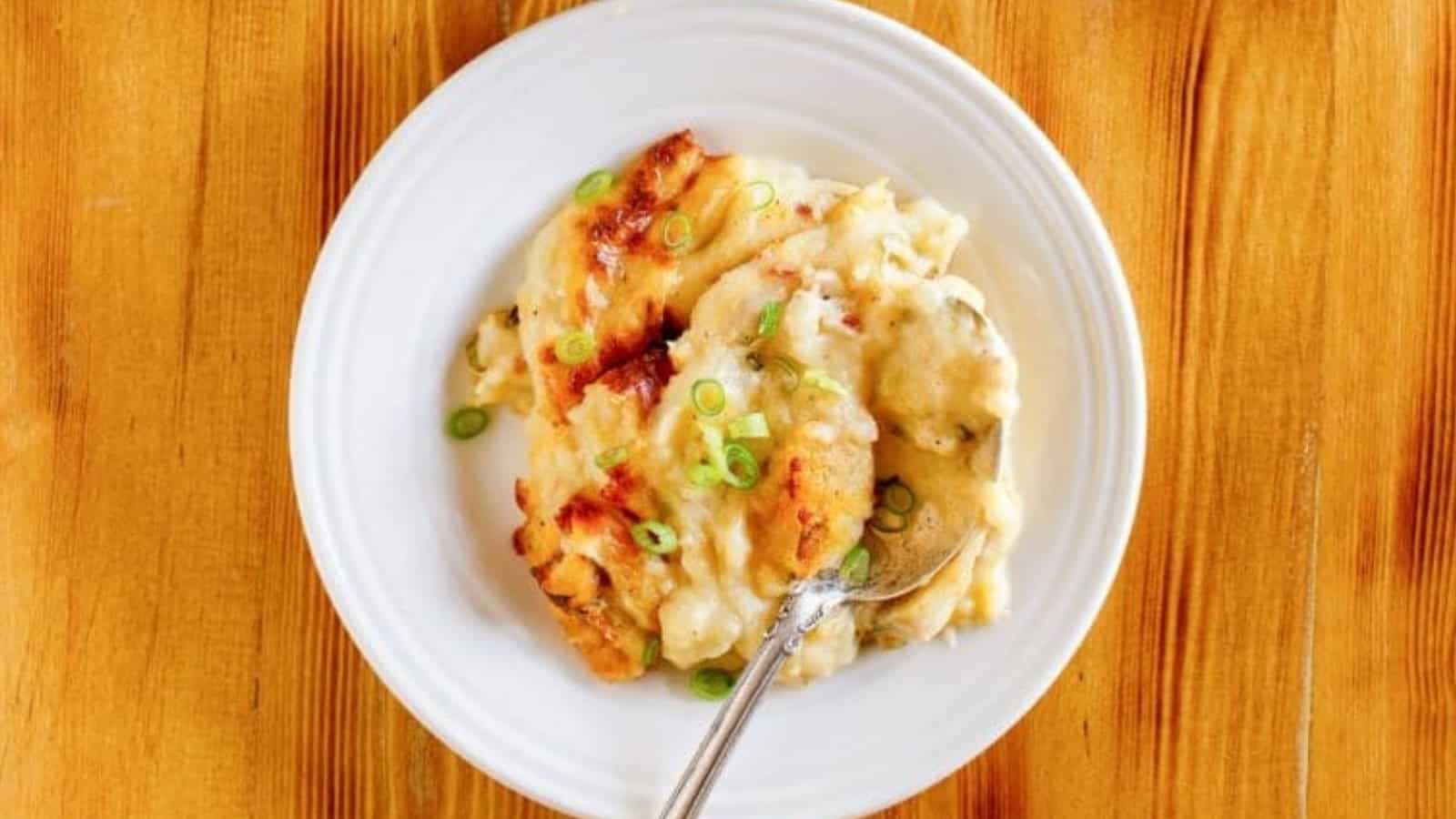 A plate of cheesy mashed potatoes topped with sliced green onions on a wooden table, with a spoon.