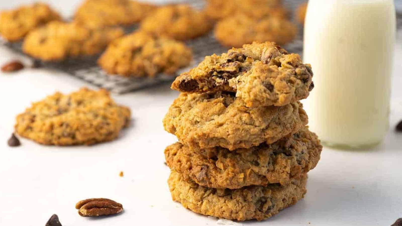 A stack of oatmeal cookies next to a glass bottle of milk, with more cookies cooling on a rack behind.