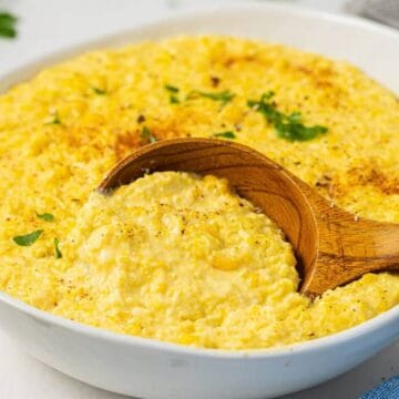 A bowl of creamy yellow corn pudding, garnished with herbs, with a wooden spoon scooping some out—perfect for your Memorial Day side dishes.