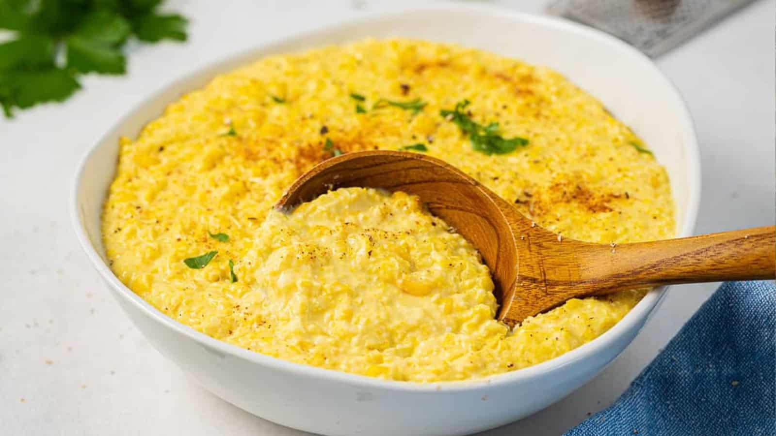 A bowl of creamy yellow corn pudding, garnished with herbs, with a wooden spoon scooping some out—perfect for your Memorial Day side dishes.