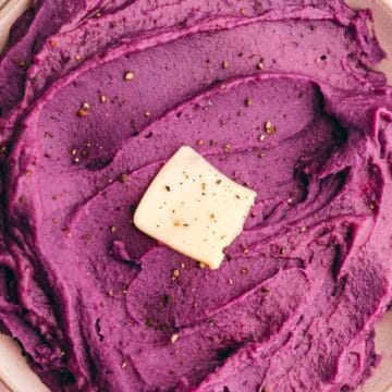 Bowl of purple mashed potatoes topped with a pat of butter and black pepper, with a wooden spoon on the side.