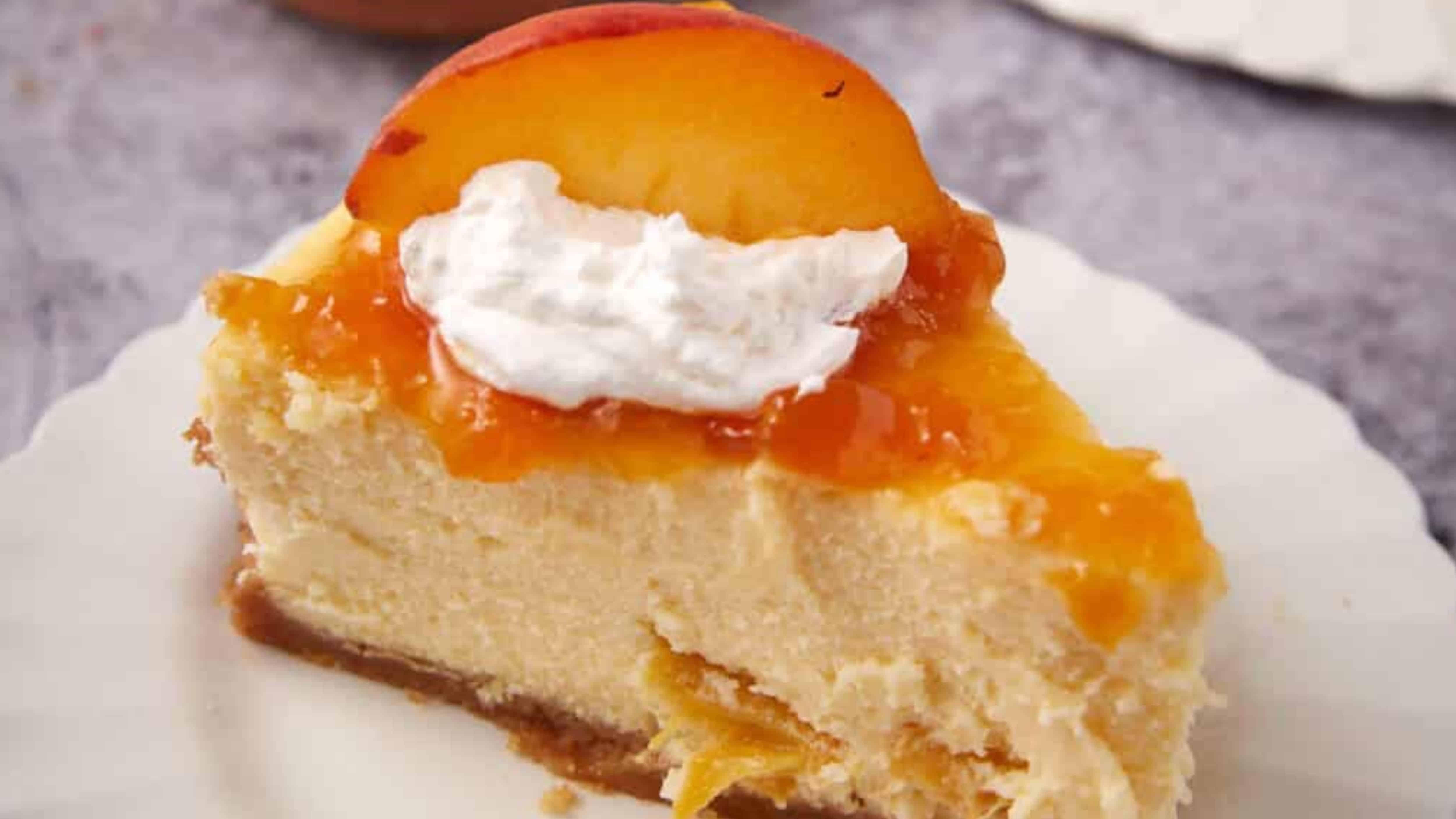 Slice of cheesecake topped with peach, whipped cream, and glaze on a white plate.