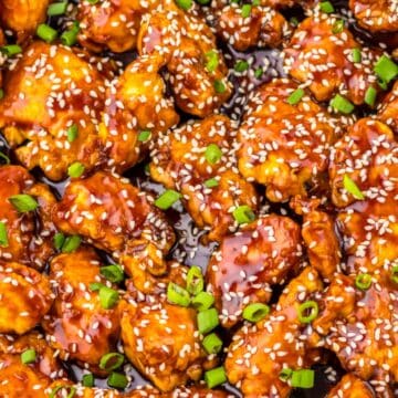 Close-up of saucy sesame chicken pieces topped with sesame seeds and chopped green onions.