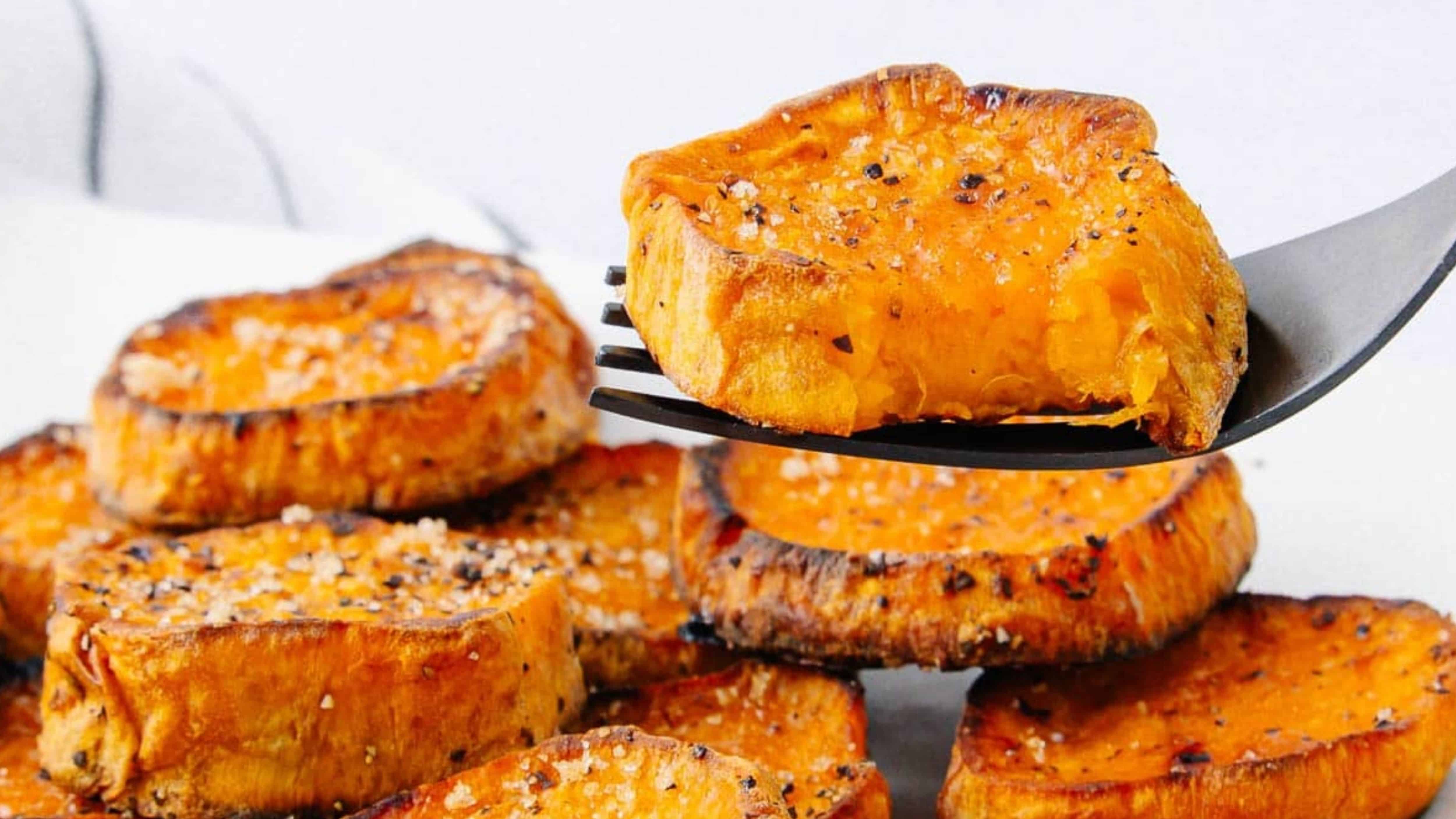 A fork holds a slice of roasted sweet potato, seasoned with salt and pepper, above a pile of more slices.