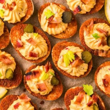 Deviled eggs served on crispy bases, topped with bacon, pickles, herbs, and garnished with lemon wedges.
