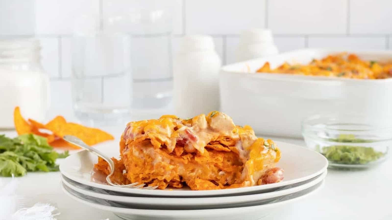 A cheesy Doritos casserole served on a plate with a fork, garnished with herbs, with a casserole dish in the background.