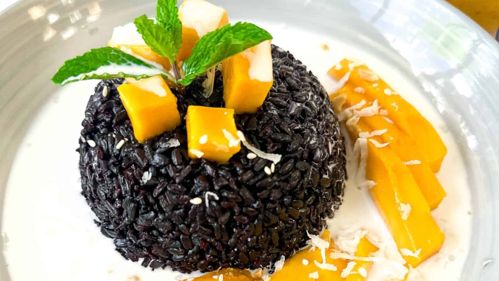 Black sticky rice dessert topped with mango chunks and mint, served with coconut milk and sliced mango on the side.