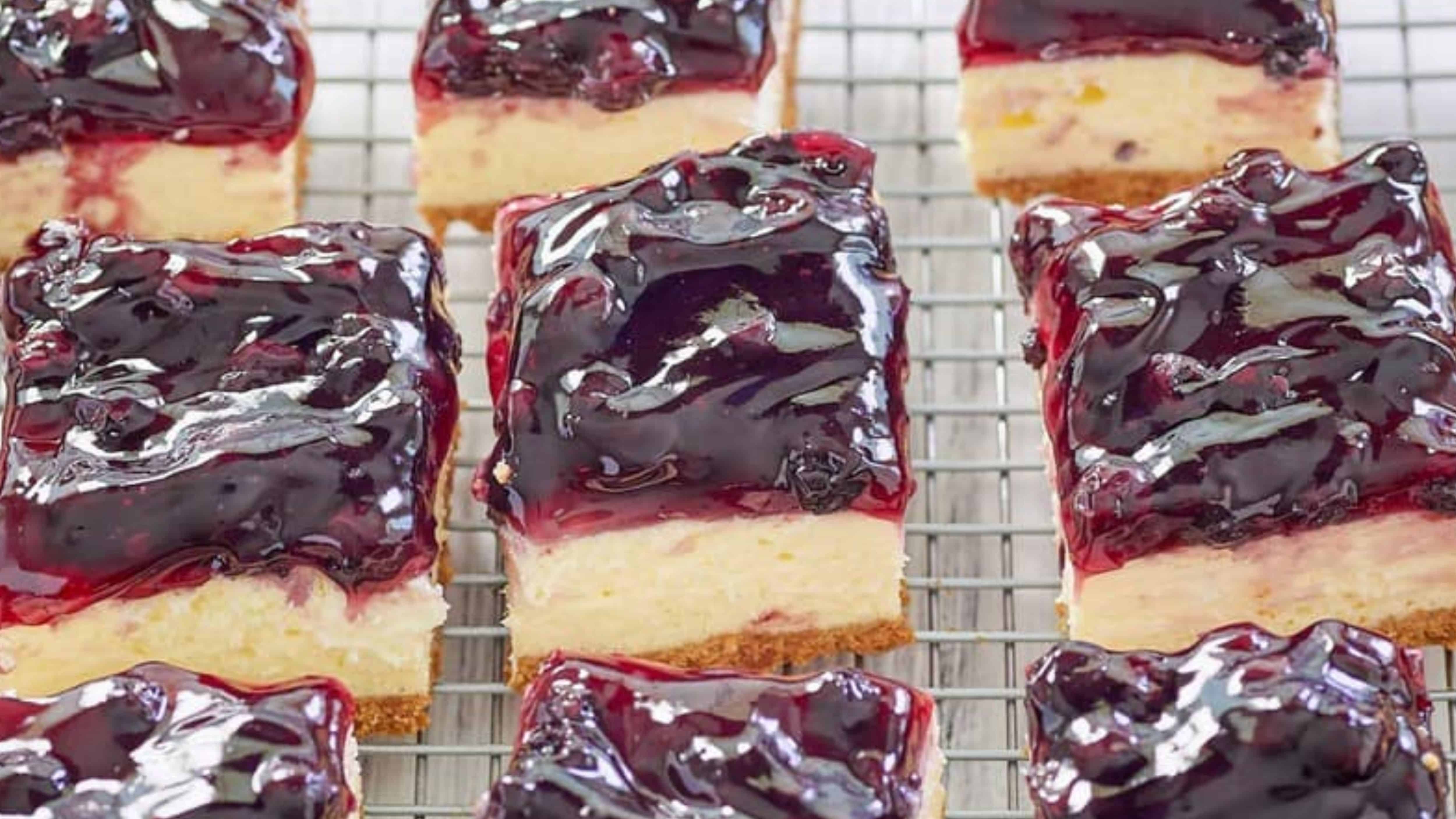Nine blueberry cheesecake bars with a shiny berry topping are cooling on a wire rack.