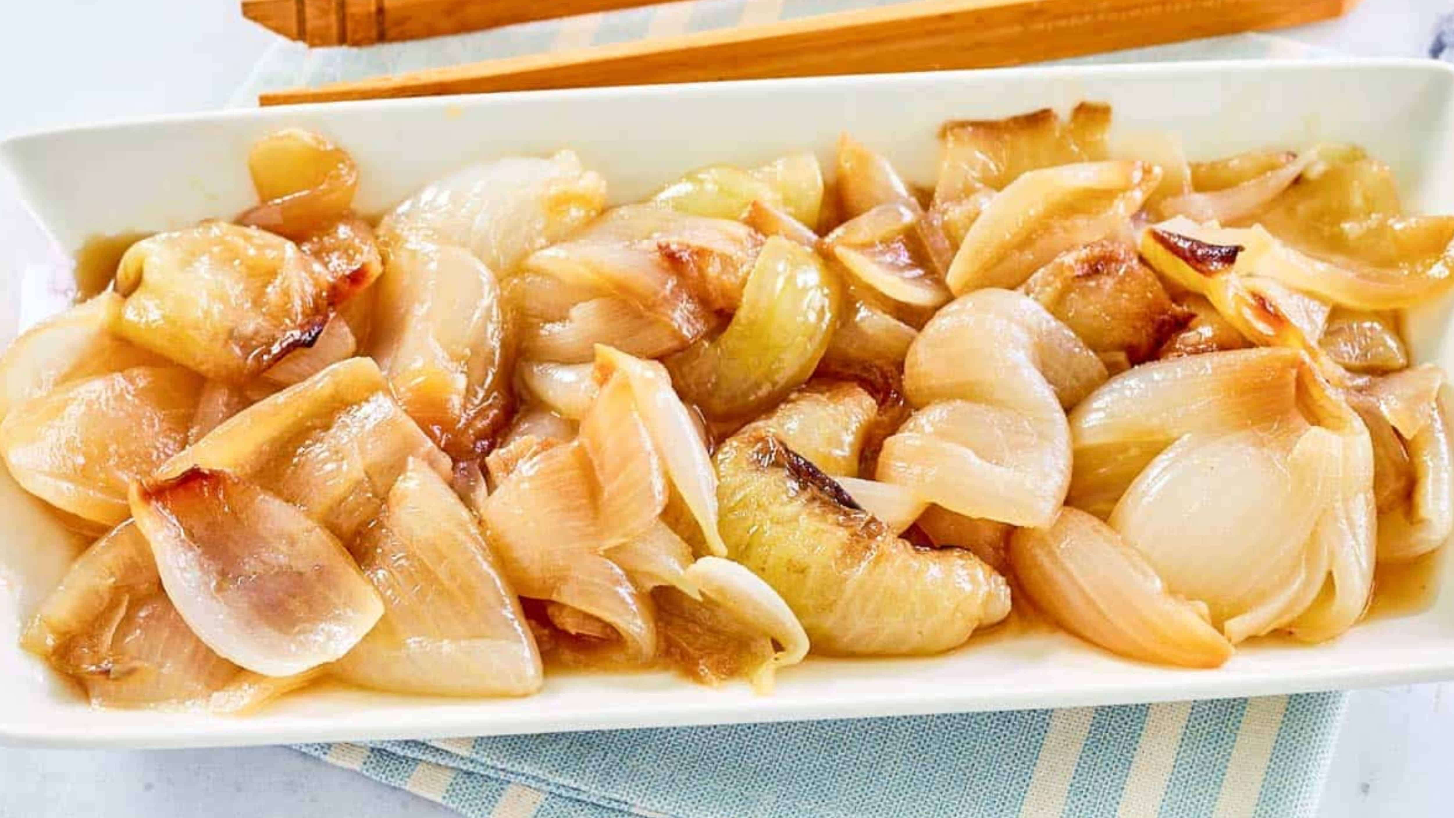 A rectangular plate of glazed, roasted onions, with chopsticks in the background on a striped cloth.