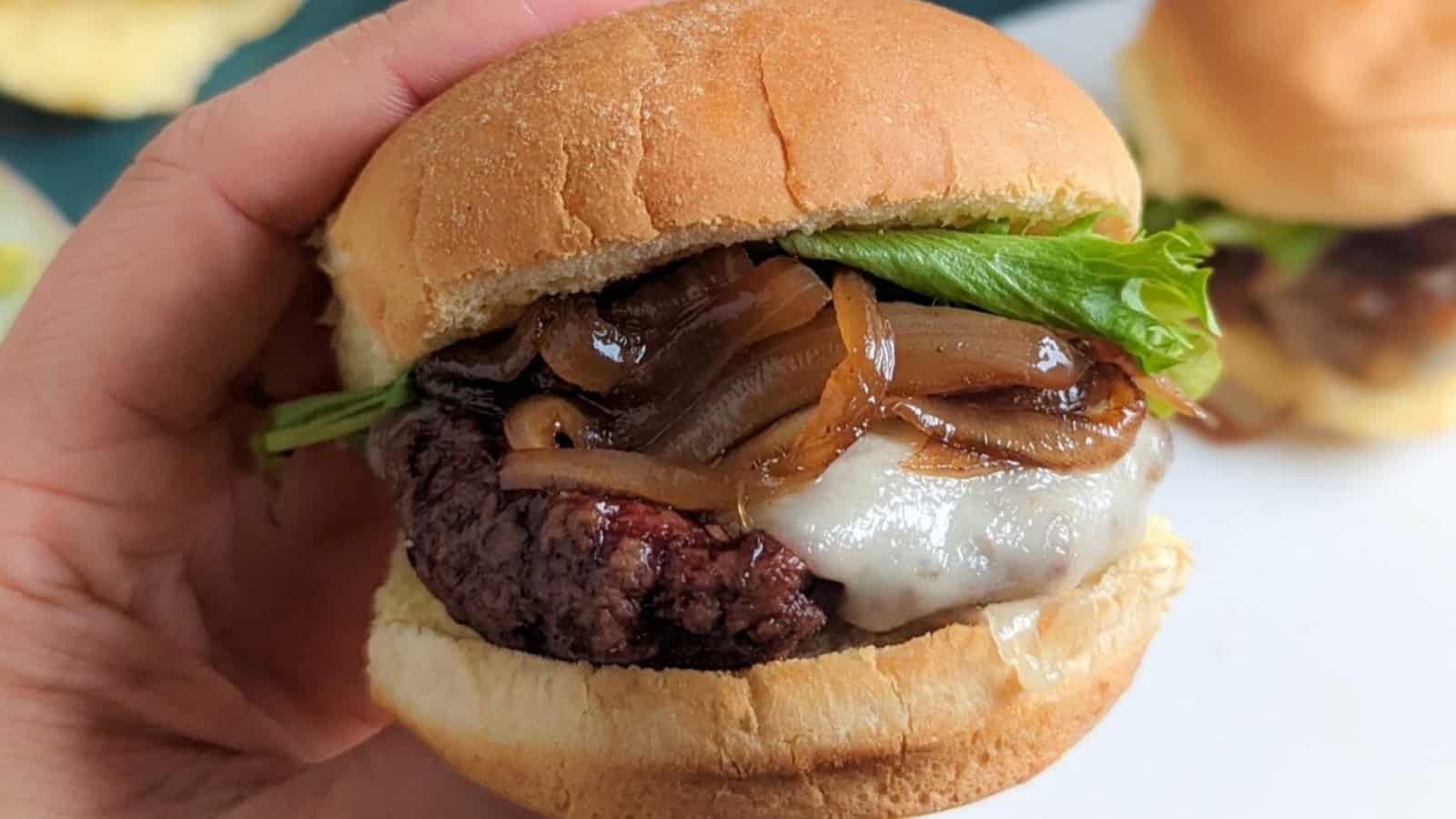 A hand holding a cheeseburger with grilled onions, lettuce, and a toasted bun.