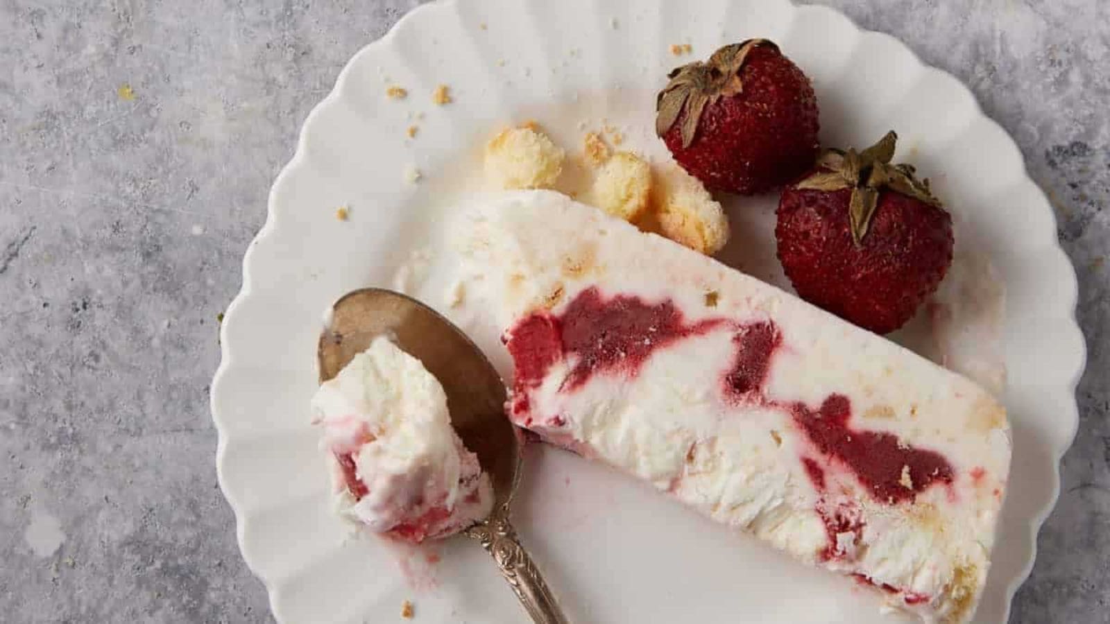 A slice of strawberry and cream dessert on a white plate with two strawberries and a spoon.