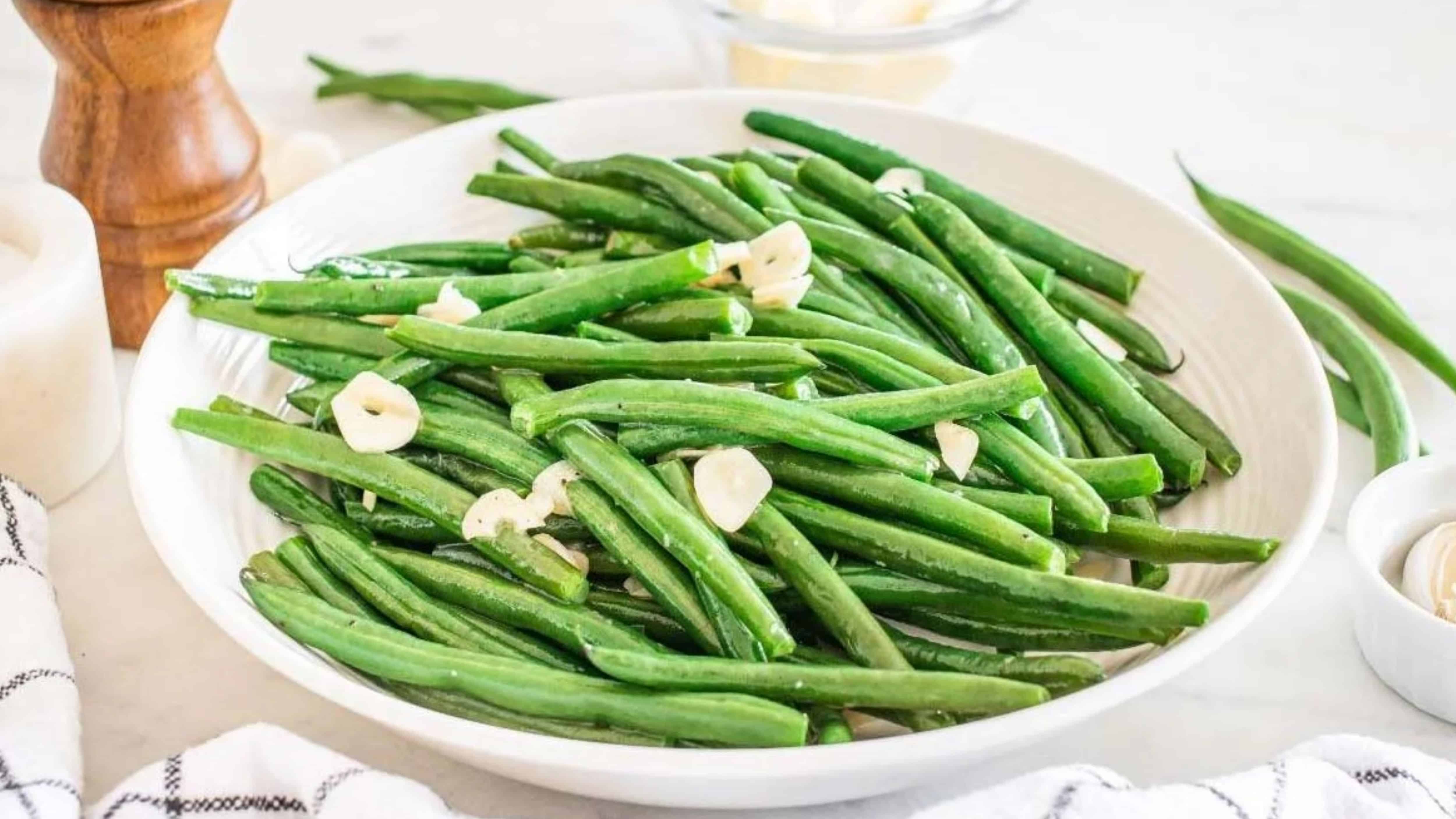 A plate of steamed green beans garnished with sliced garlic on a white table.