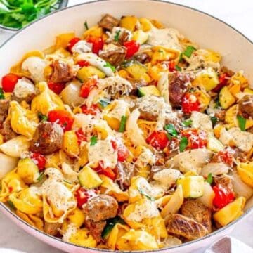 A skillet filled with tortellini, beef, cherry tomatoes, zucchini, and a creamy sauce, garnished with herbs.