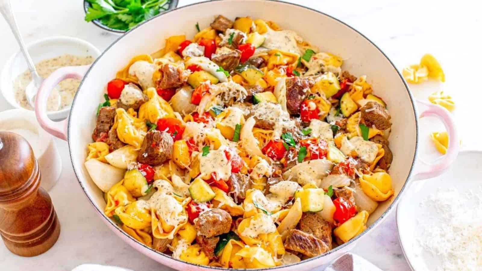 A skillet filled with tortellini, beef, cherry tomatoes, zucchini, and a creamy sauce, garnished with herbs.