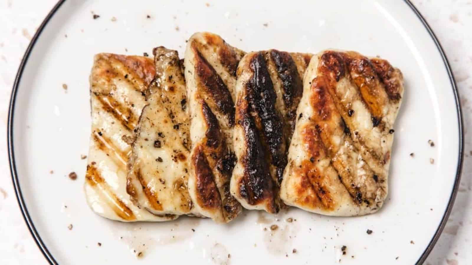 Five grilled halloumi cheese slices with grill marks on a white plate, seasoned with black pepper.