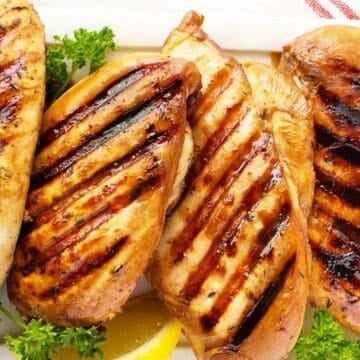 Grilled chicken breasts with char marks, garnished with parsley and lemon wedges on a white plate.