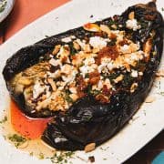 Charred eggplant topped with feta cheese, herbs, almonds, and chili oil on a white plate.