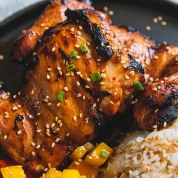 Grilled chicken with sesame seeds, served with white rice and colorful diced vegetables on a black plate.