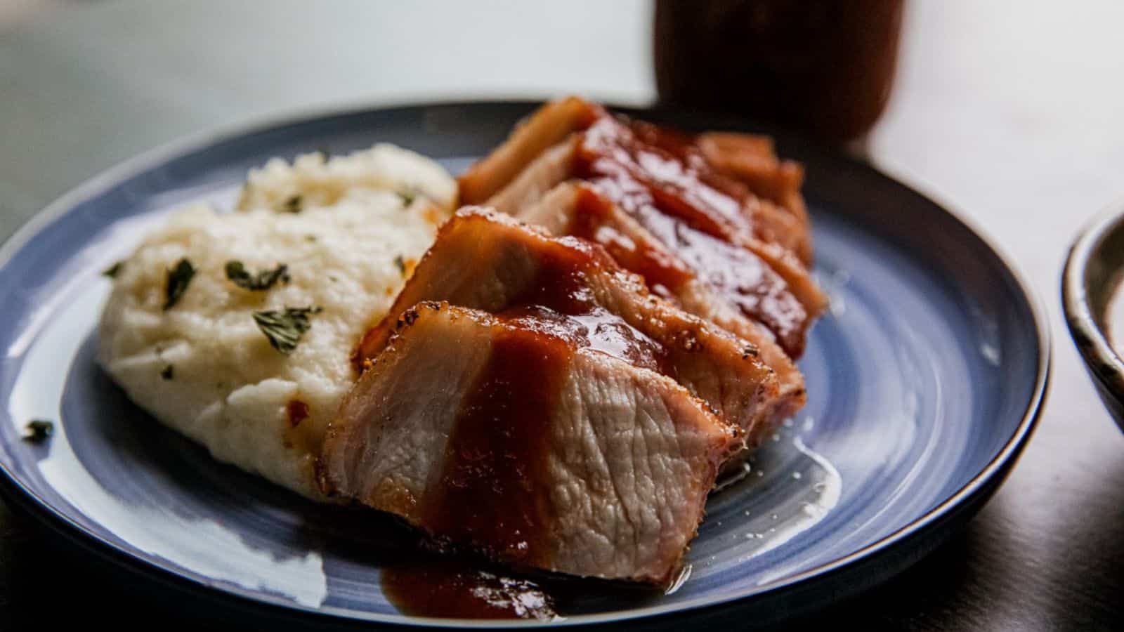 Sliced pork with barbecue sauce and mashed potatoes garnished with herbs on a blue plate.