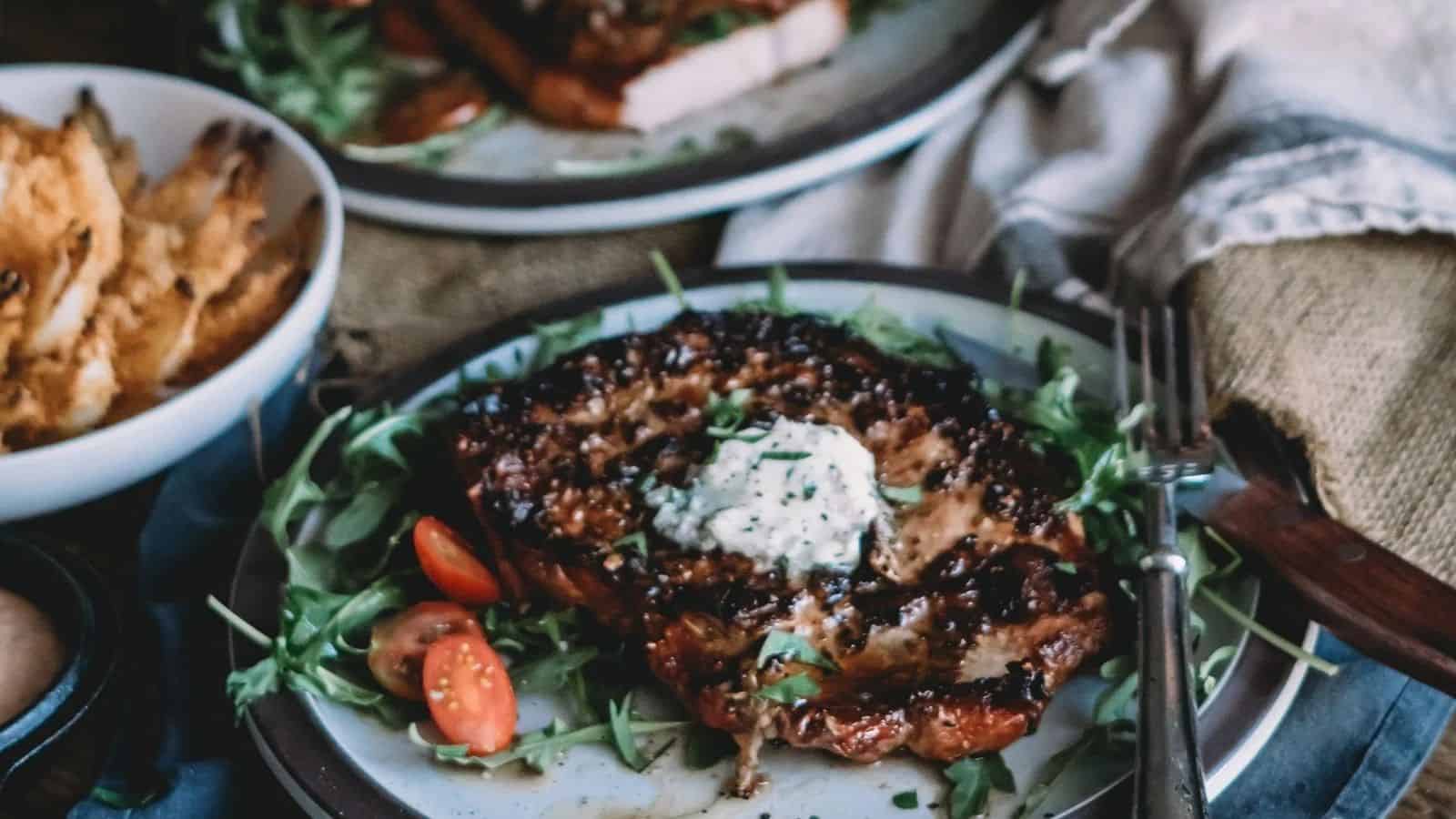 Grilled steak topped with herb butter, cherry tomatoes, and greens on a plate with a fork.