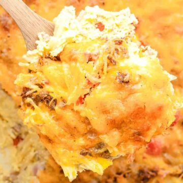 A wooden spoon holds a cheesy, golden-brown casserole with visible chunks of vegetables and melted cheese.