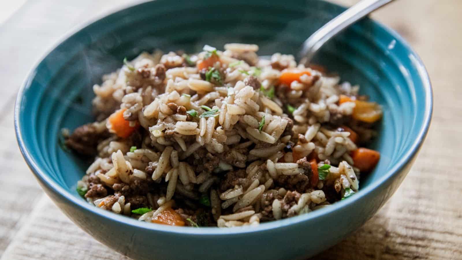 A blue bowl of rice mixed with ground meat, carrots, and herbs, with a spoon inside the bowl.