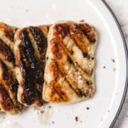 Three pieces of grilled halloumi cheese with grill marks on a white plate, sprinkled with black pepper.