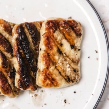 Three pieces of grilled halloumi cheese with grill marks on a white plate, sprinkled with black pepper.
