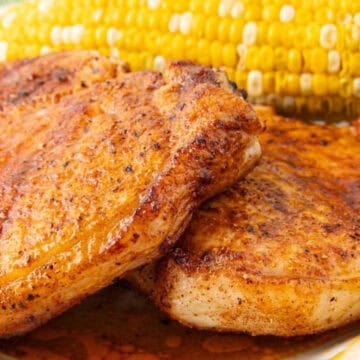 Two seasoned pork chops on a plate with a serving of corn on the cob and a sprig of parsley.