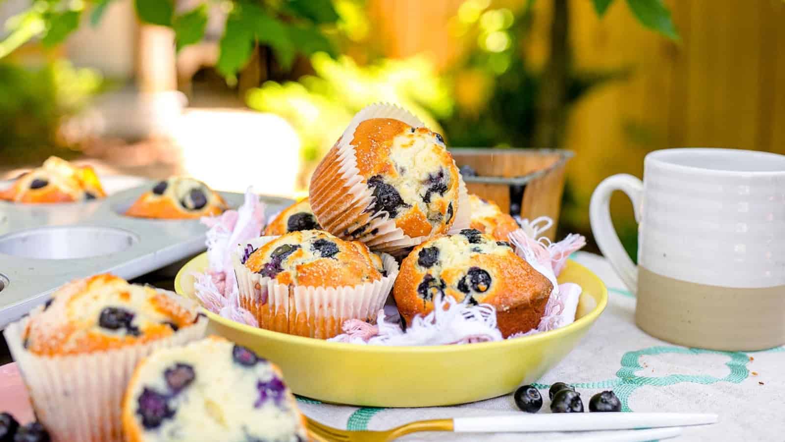 A bowl of blueberry muffins, a delicious addition to berry desserts, sits on a table beside a white mug, with greenery in the background.