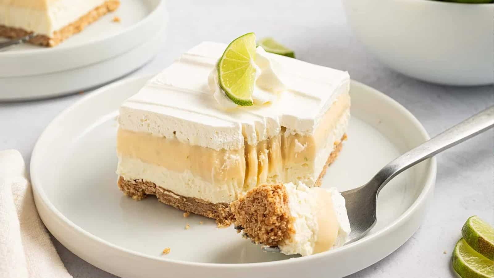 A creamy dessert bar with layers, topped with whipped cream and a lime slice, on a white plate with a fork.