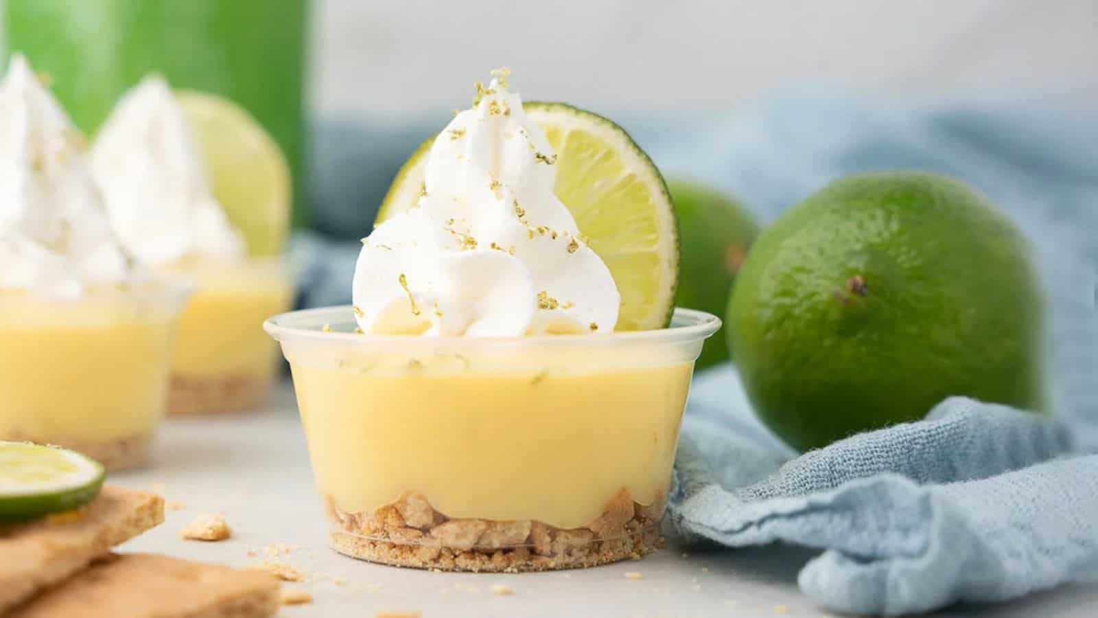 Mini key lime pie in a clear cup topped with whipped cream, with fresh limes and a blue cloth in the background.