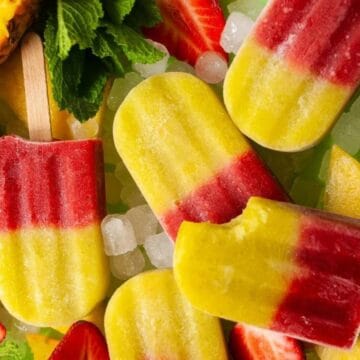 Assorted red and yellow fruit popsicles with ice, strawberries, and pineapple slices on a green background.