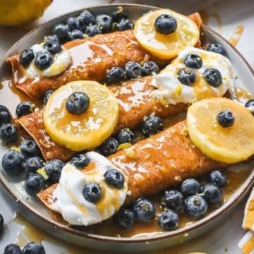 Three crepes topped with whipped cream, blueberries, lemon slices, and syrup on a plate.