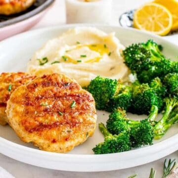 A plate with chicken patties, mashed potatoes with olive oil, and roasted broccoli next to lemon slices.