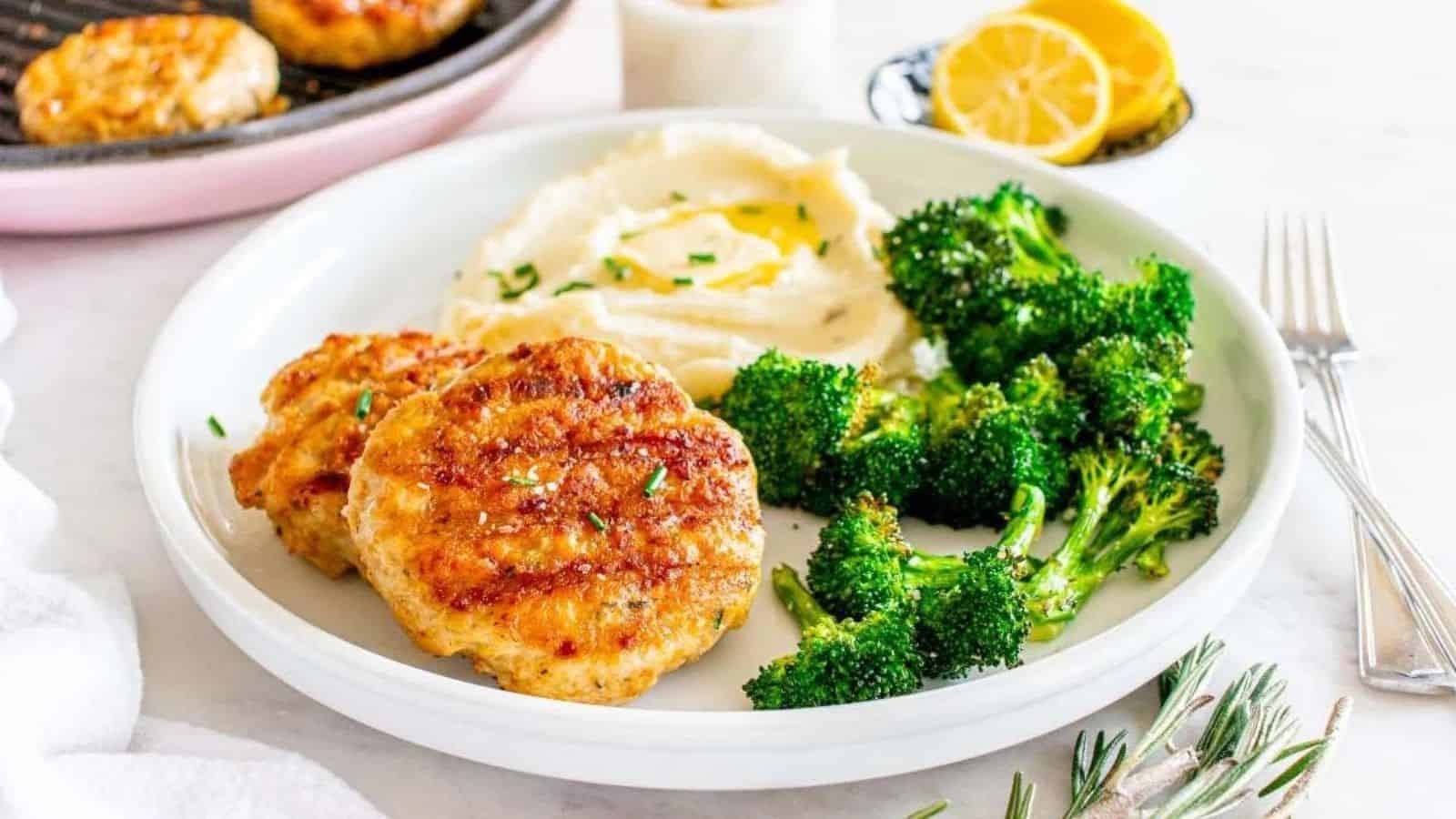 A plate with chicken patties, mashed potatoes with olive oil, and roasted broccoli next to lemon slices.