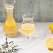 A glass of pale yellow cocktail on a marble surface with a carafe, lemon, and lavender sprigs nearby.