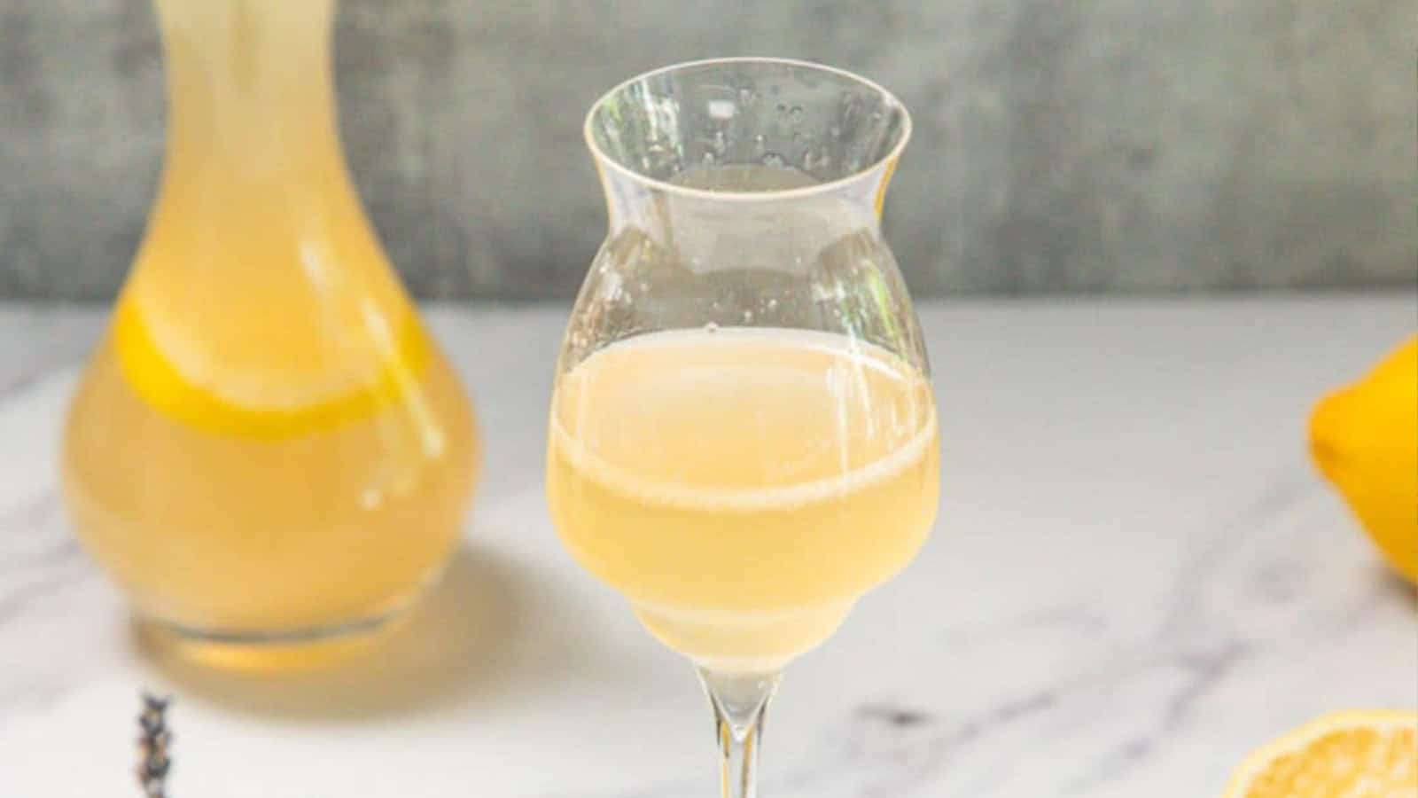 A glass of pale yellow drink on a white surface, with a carafe and lemon in the background.