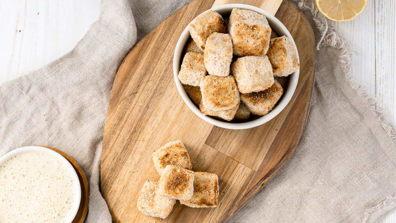 Bowl of sugar cubes on a wooden board with a beige cloth and a frothy drink nearby.