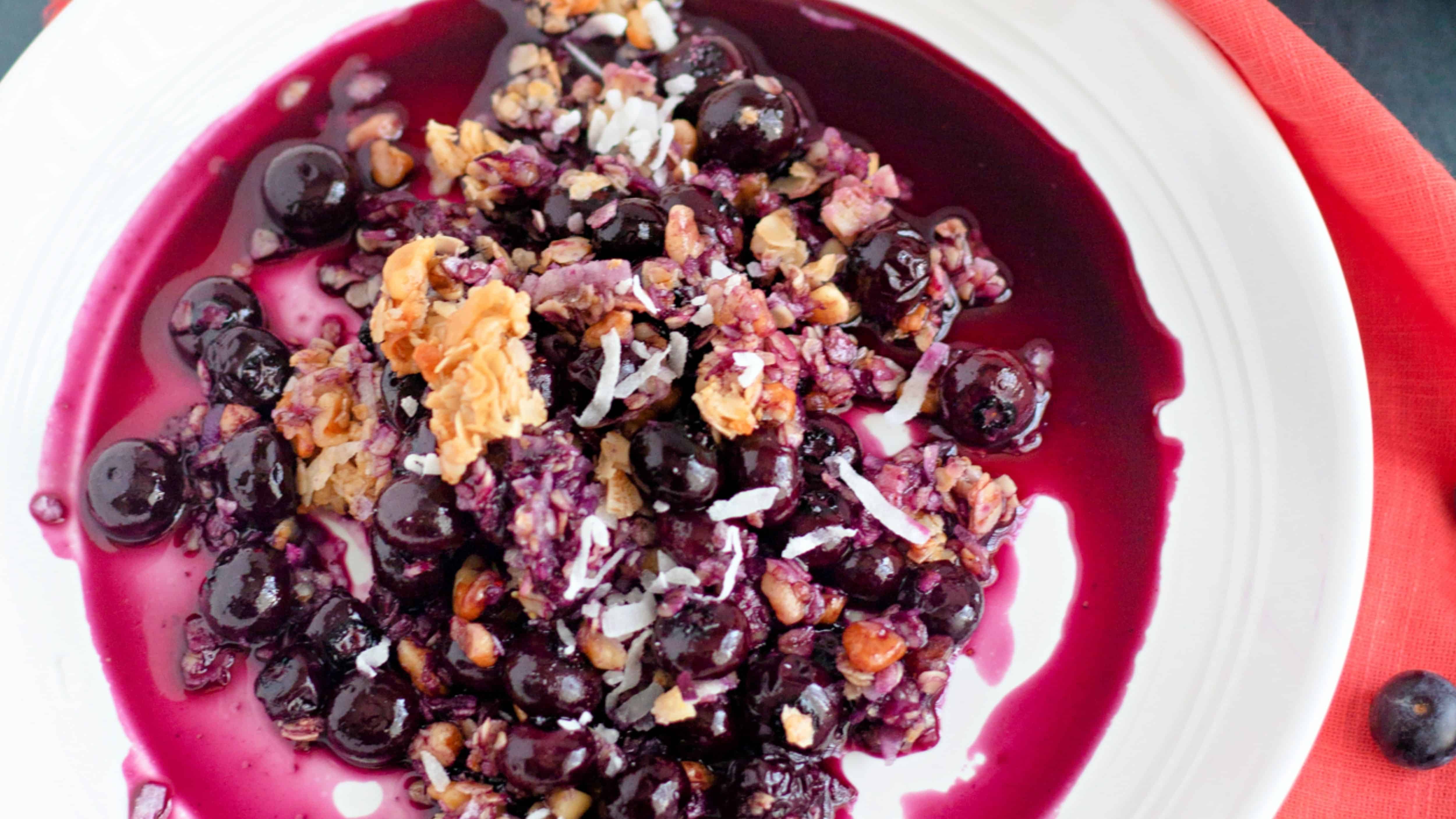 A close-up of blueberry crisp with oats, nuts, and syrup on a white plate with an orange napkin nearby.