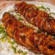 Caramelized sweet potato slices rest on a bed of creamy yogurt, topped with green onions and vibrant red spices on a white plate.