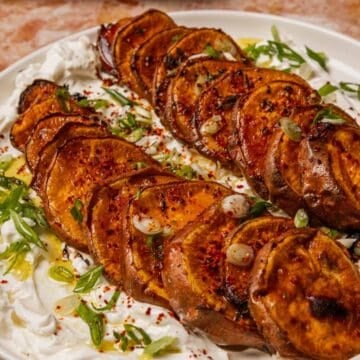 Caramelized sweet potato slices rest on a bed of creamy yogurt, topped with green onions and vibrant red spices on a white plate.