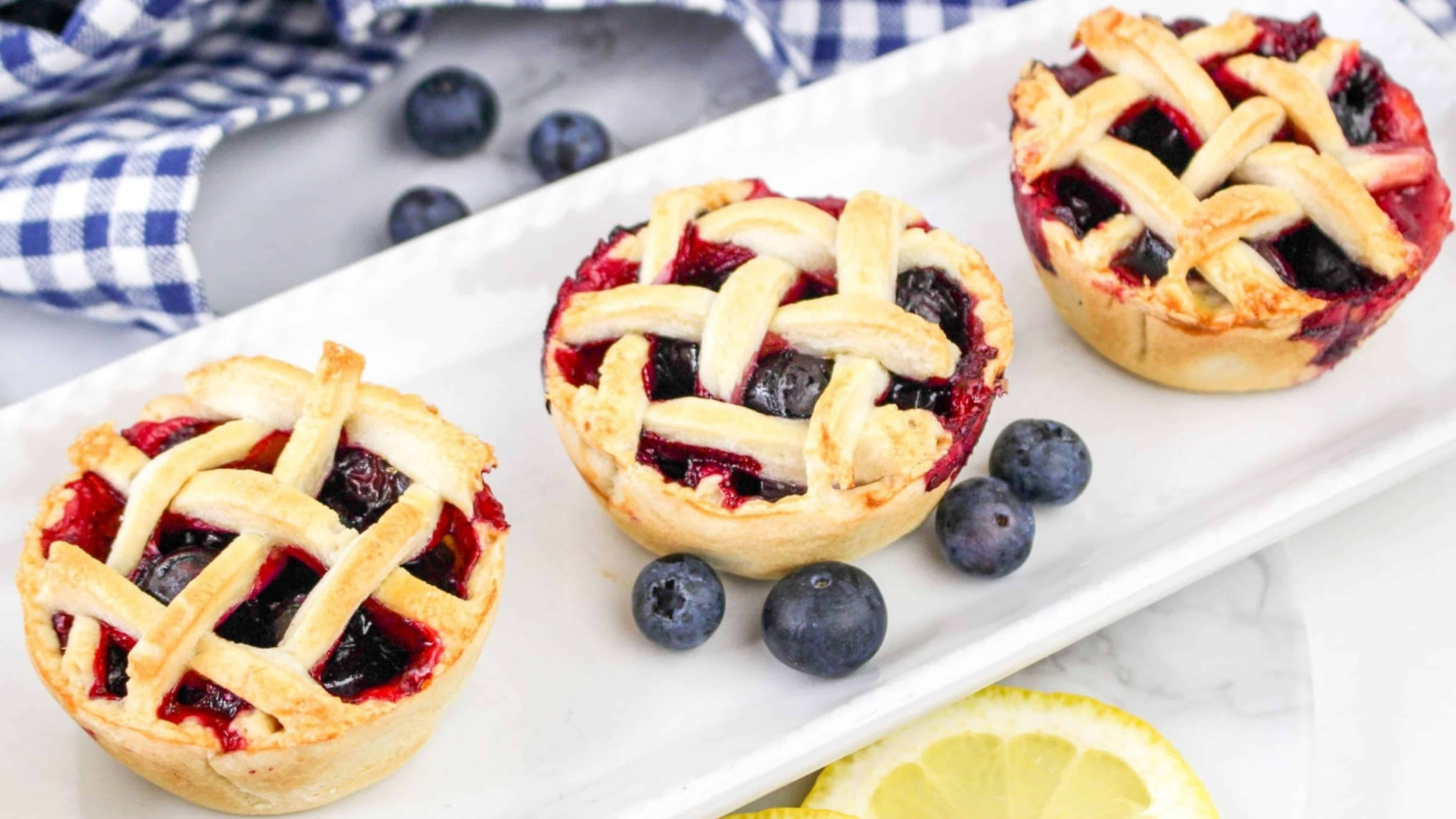 Three mini blueberry pies with lattice tops on a rectangular white plate, surrounded by fresh blueberries and lemon slices, and a blue checkered cloth in the background.